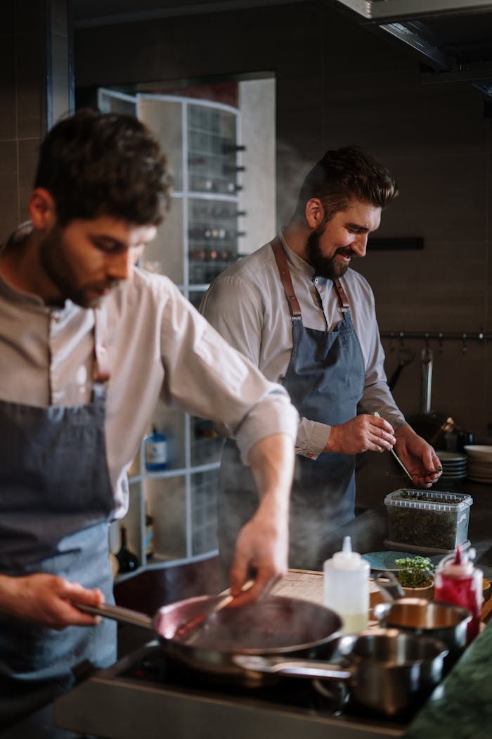 Two male chefs wearing aprons cooking together in a professional kitchen setting.